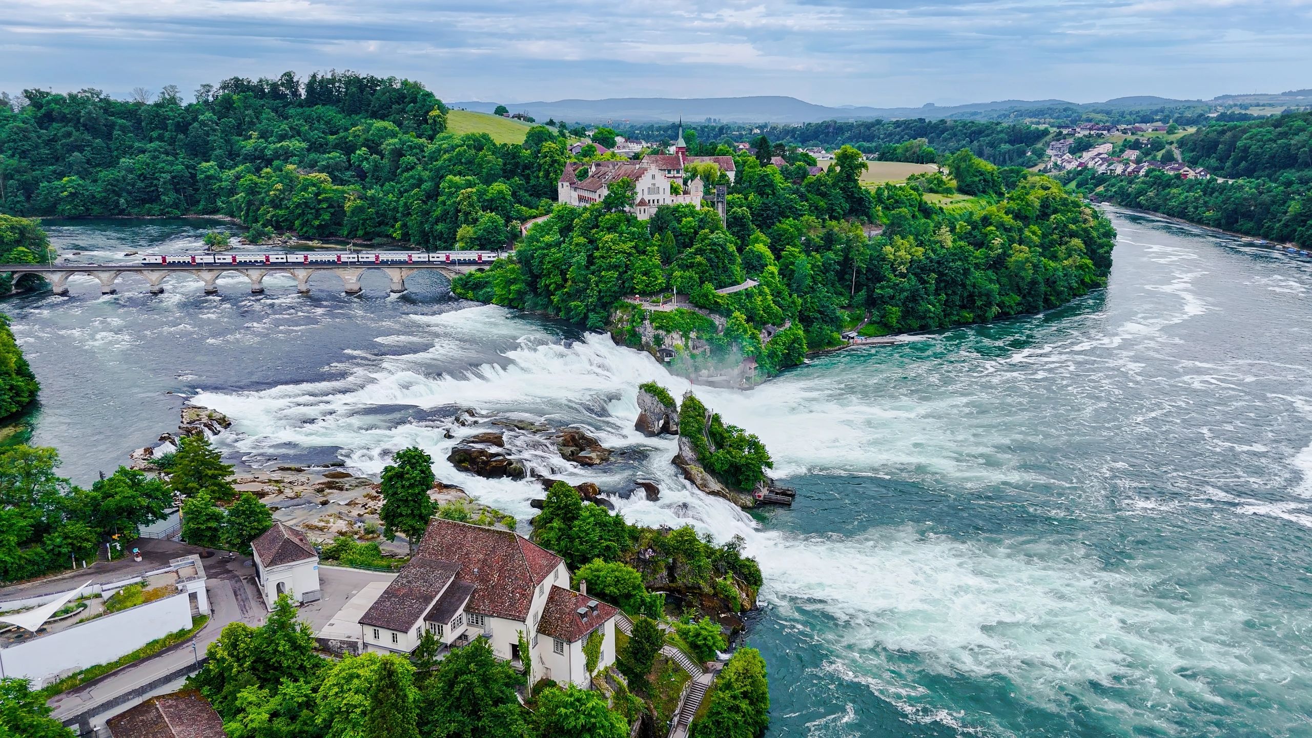 Aerial view of Rhine Falls with train crossing historic railway bridge, Laufen Castle on green hilltop, lush forest surrounding powerful waterfall on the Rhine River, Swiss Alps travel and transportation concept